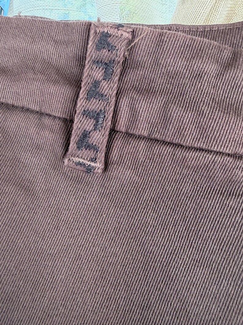 May include: Close-up of brown fabric with a textured weave. A belt loop is visible, featuring the embroidered word "AVA" in dark thread. The fabric appears to be a sturdy material, possibly denim or a similar textile.