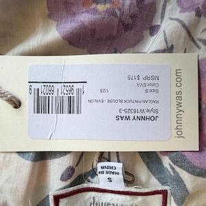 May include: A close-up of a floral print blouse with a tag. The tag reads "JOHNNY WAS" and "MADE IN CHINA". The blouse has a cream background with purple and orange floral patterns. The tag also shows the size as "S".