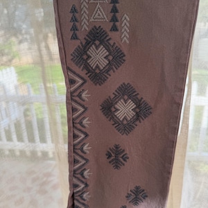 May include: Close-up of mauve-coloured trouser leg with embroidered geometric patterns in shades of grey and off-white. The design includes triangles, diamonds, and zigzags, creating a textured, decorative look. The trousers are made of a soft fabric.