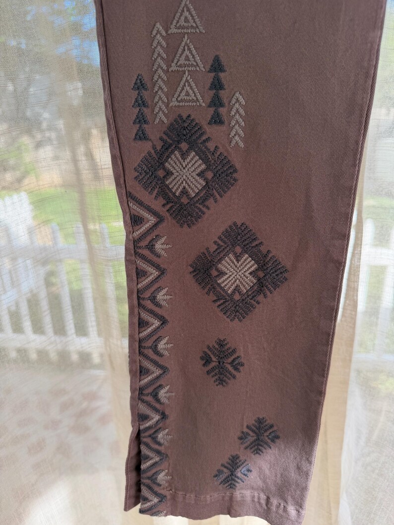May include: Close-up of mauve-coloured trouser leg with embroidered geometric patterns in shades of grey and off-white. The design includes triangles, diamonds, and zigzags, creating a textured, decorative look. The trousers are made of a soft fabric.