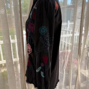 以下が含まれることがあります： カラフルな花の刺繍が施された黒いボタンダウンシャツ。長袖シャツは、ピンク、赤、青、緑の色合いで、ほつれた裾と複雑なステッチが特徴です。シャツはマネキンに展示されています。