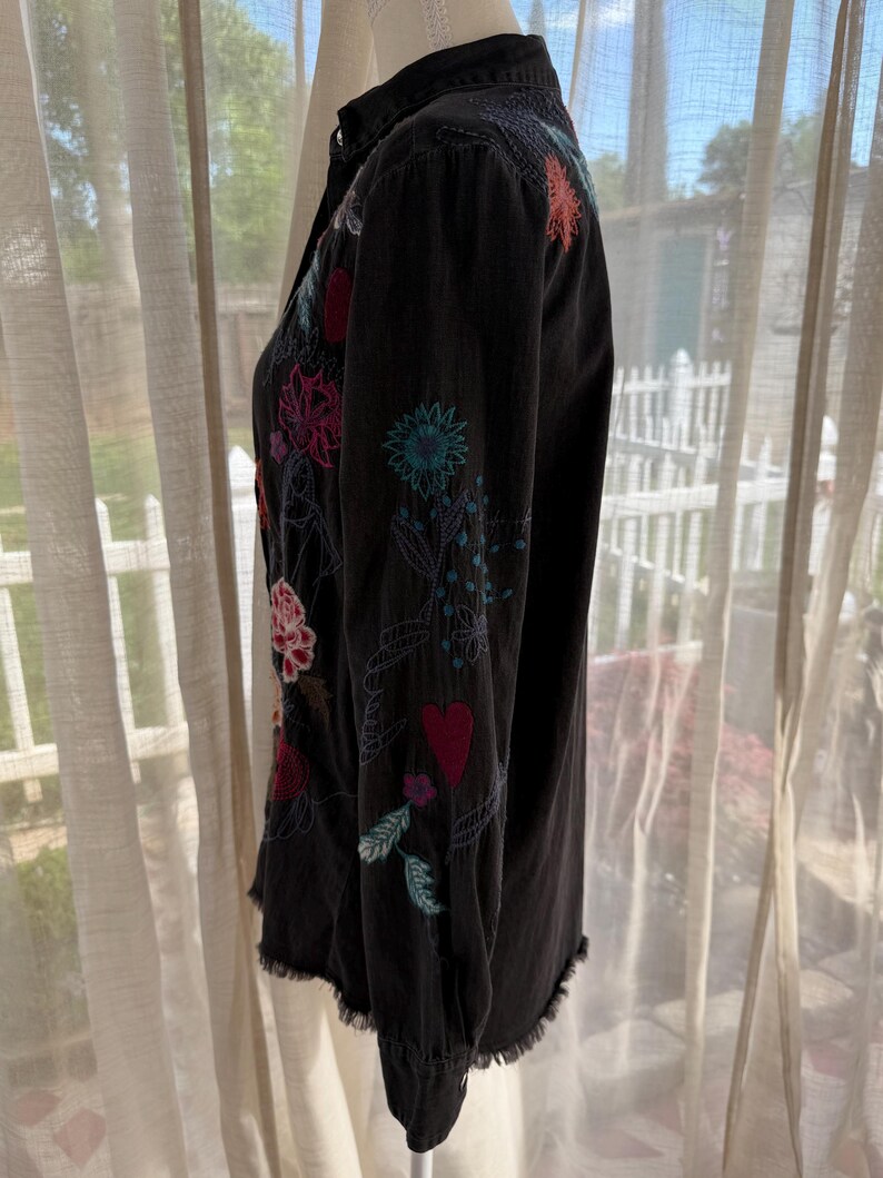 以下が含まれることがあります： カラフルな花の刺繍が施された黒いボタンダウンシャツ。長袖シャツは、ピンク、赤、青、緑の色合いで、ほつれた裾と複雑なステッチが特徴です。シャツはマネキンに展示されています。