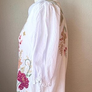 May include: White embroidered blouse with a collared neckline and three-quarter sleeves. The floral embroidery features shades of pink, gold, and green. The blouse is displayed on a mannequin.