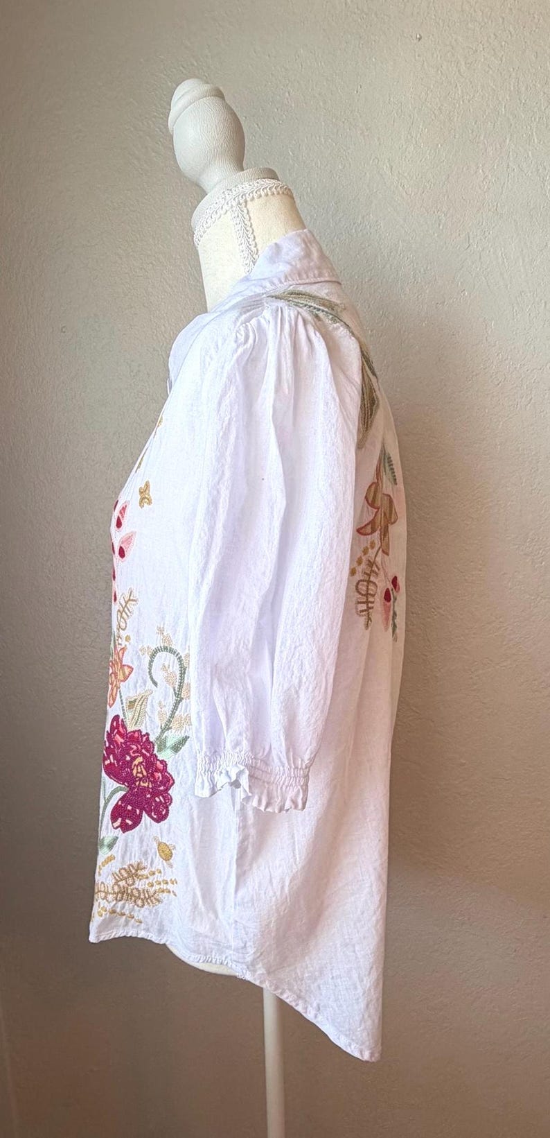 May include: White embroidered blouse with a collared neckline and three-quarter sleeves. The floral embroidery features shades of pink, gold, and green. The blouse is displayed on a mannequin.