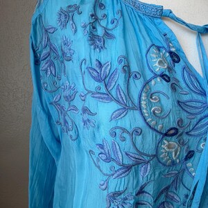 Puede incluir: Una blusa azul claro con mangas largas y bordados florales intrincados en tonos azules. La blusa presenta un cuello en V con lazos y costuras delicadas a lo largo del escote y los hombros. La tela parece ligera y ligeramente texturizada.