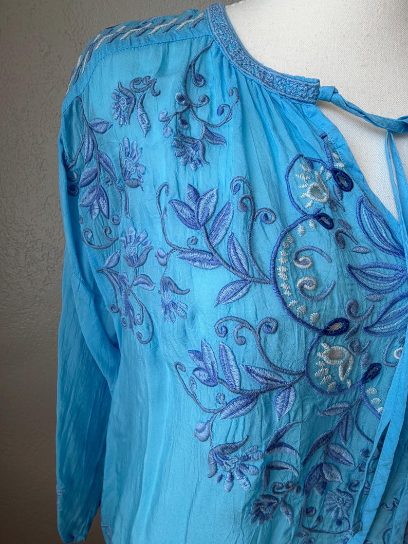 Puede incluir: Una blusa azul claro con mangas largas y bordados florales intrincados en tonos azules. La blusa presenta un cuello en V con lazos y costuras delicadas a lo largo del escote y los hombros. La tela parece ligera y ligeramente texturizada.