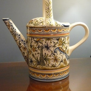 May include: A white ceramic watering can with a floral design in shades of blue, yellow, and green. The handle is shaped like a leaf and has a green and white design.