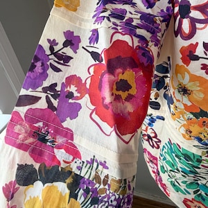May include: A cream-colored garment with a vibrant floral pattern. The design features large, colorful flowers in shades of red, purple, yellow, and orange. The fabric appears to be lightweight, with visible seams and a flowing silhouette.
