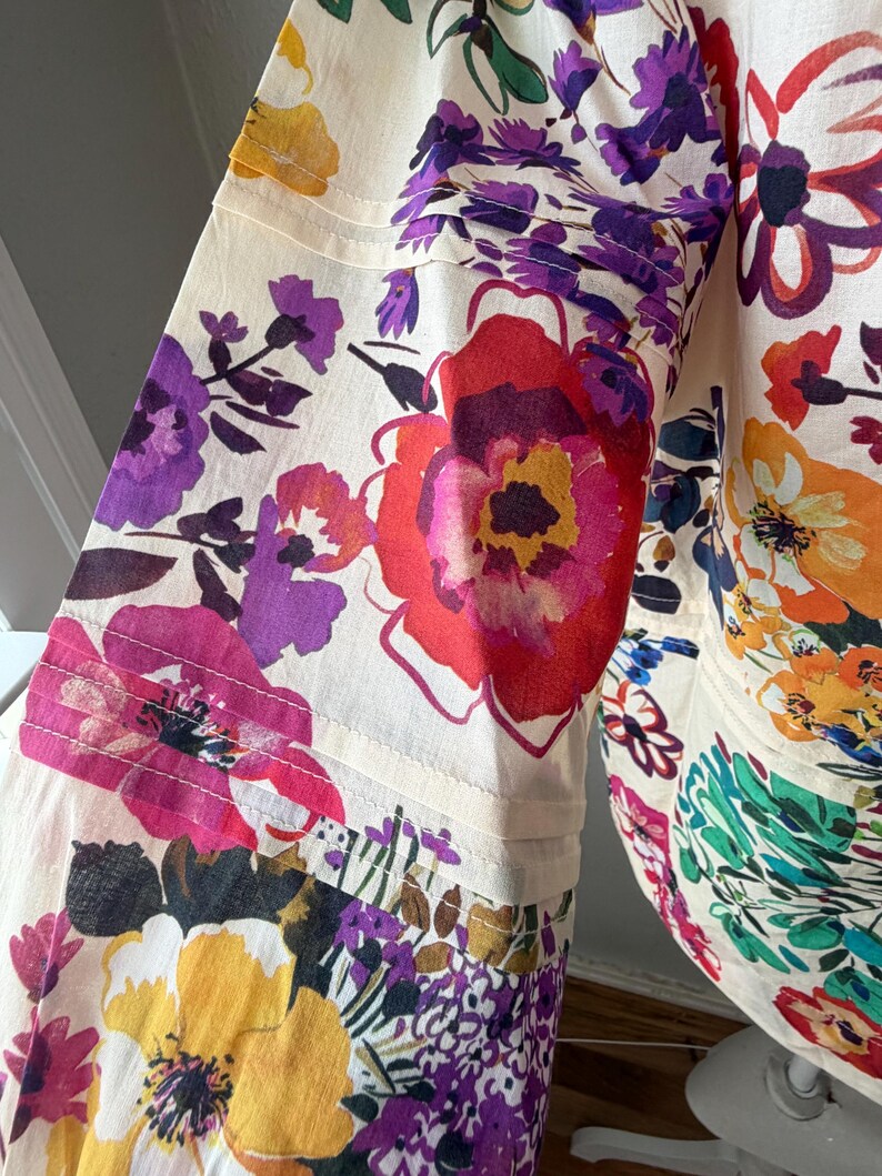 May include: A cream-colored garment with a vibrant floral pattern. The design features large, colorful flowers in shades of red, purple, yellow, and orange. The fabric appears to be lightweight, with visible seams and a flowing silhouette.