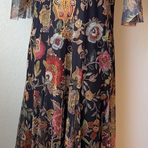 May include: Black floral print dress with sheer sleeves and a decorative gold embroidered neckline. The dress features a flowing skirt and a detailed floral pattern in shades of red, pink, and gold.