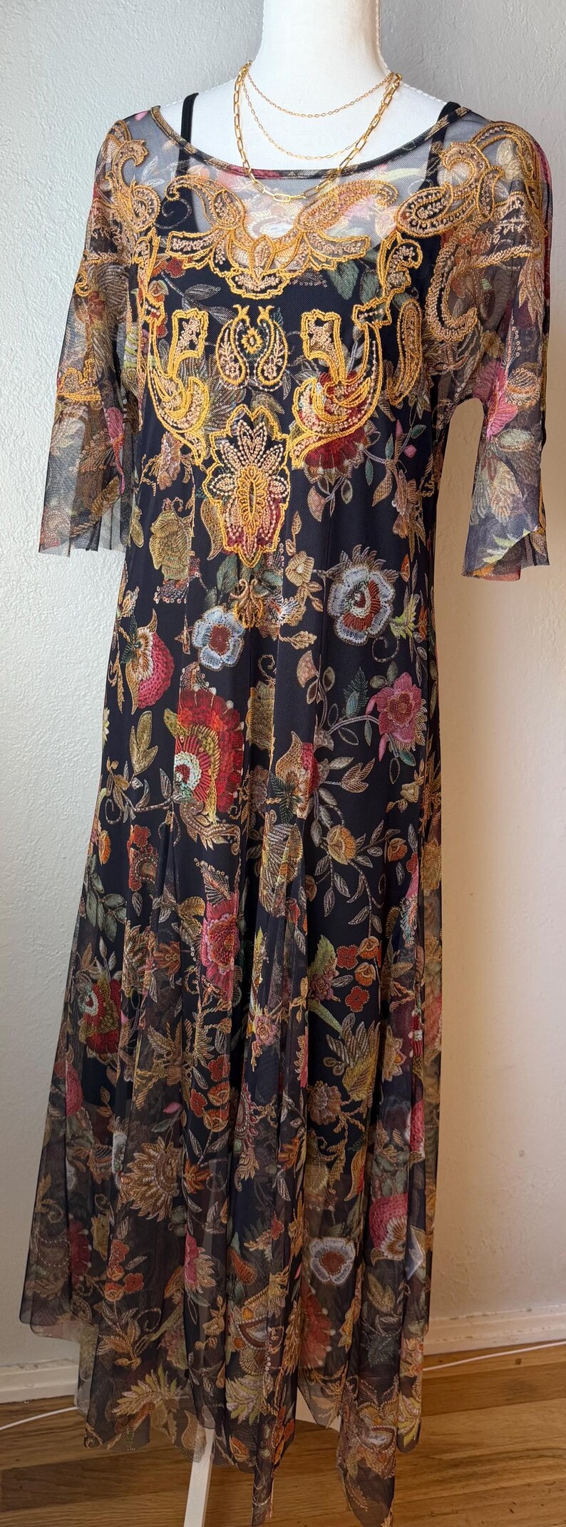 May include: Black floral print dress with sheer sleeves and a decorative gold embroidered neckline. The dress features a flowing skirt and a detailed floral pattern in shades of red, pink, and gold.