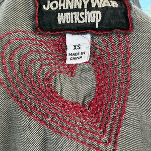 以下が含まれることがあります： 背中に赤い刺繍のハートのデザインが施されたグレーのデニムジャケットのクローズアップ。 ジャケットには、黒いパッチに「JOHNNY WAS WORKSHOP」の文字が書かれています。 XSサイズのタグが見え、「MADE IN CHINA」と記載されています。