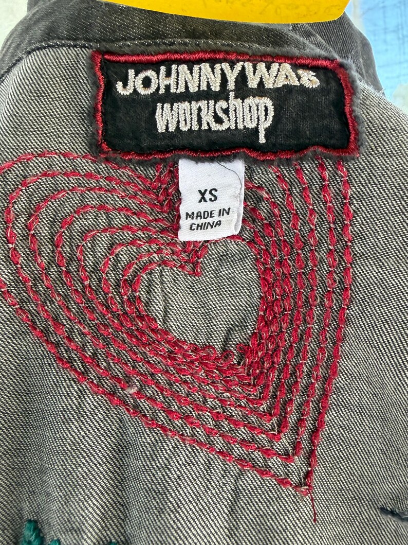 以下が含まれることがあります： 背中に赤い刺繍のハートのデザインが施されたグレーのデニムジャケットのクローズアップ。 ジャケットには、黒いパッチに「JOHNNY WAS WORKSHOP」の文字が書かれています。 XSサイズのタグが見え、「MADE IN CHINA」と記載されています。