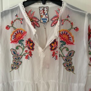 May include: White embroidered tunic with colorful floral designs in red, pink, yellow, and blue. The tunic features a V-neckline with a drawstring and a tag that reads "Johnny Was". The sleeves are long and the fabric appears to be lightweight.