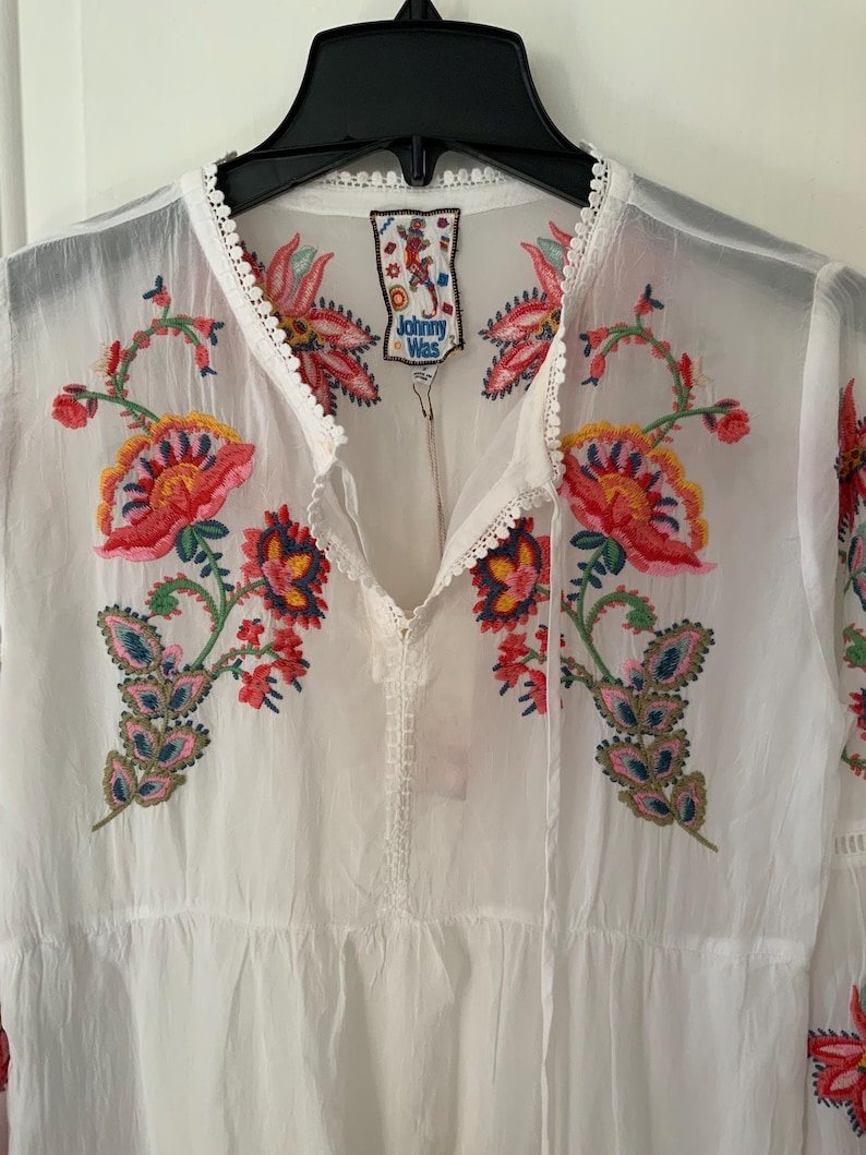 May include: White embroidered tunic with colorful floral designs in red, pink, yellow, and blue. The tunic features a V-neckline with a drawstring and a tag that reads "Johnny Was". The sleeves are long and the fabric appears to be lightweight.