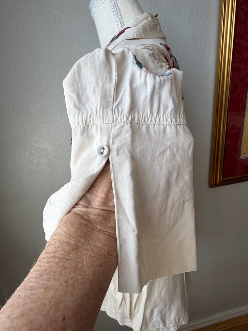 May include: Off-white button-down shirt with floral embroidery on the collar and shoulder. The shirt is displayed on a mannequin, with a buttoned cuff and a visible sleeve. The fabric appears to be cotton or a similar material.