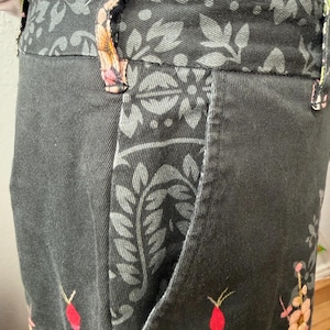 Puede incluir: Un pantal&oacute;n gris oscuro con cintura y ribetes de bolsillo con un patr&oacute;n floral en negro y gris. El pantal&oacute;n est&aacute; decorado con capullos de rosa rosas y flores amarillas bordadas en la parte inferior, incluyendo trabillas para cintur&oacute;n.