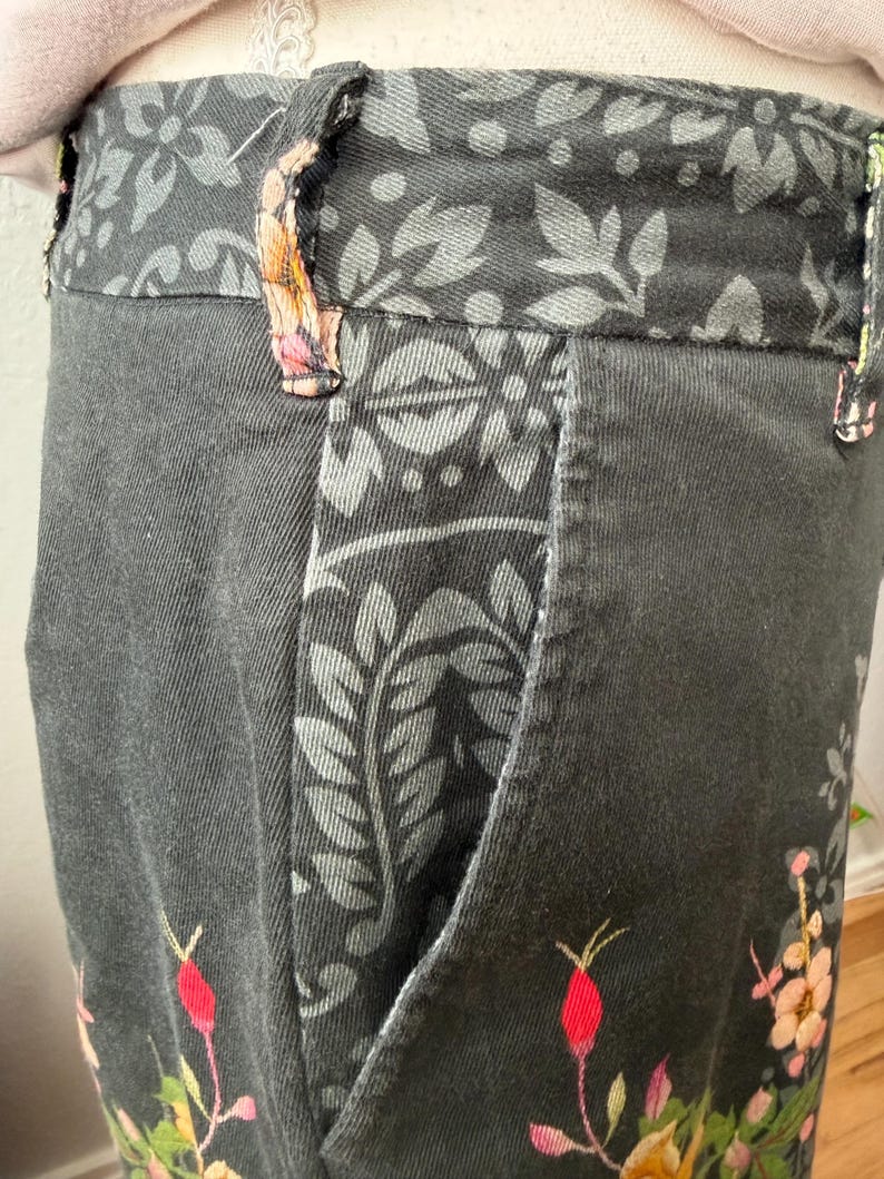 Puede incluir: Un pantal&oacute;n gris oscuro con cintura y ribetes de bolsillo con un patr&oacute;n floral en negro y gris. El pantal&oacute;n est&aacute; decorado con capullos de rosa rosas y flores amarillas bordadas en la parte inferior, incluyendo trabillas para cintur&oacute;n.