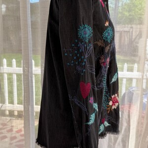 以下が含まれることがあります： カラフルな花の刺繍が施された黒のデニムシャツ。 シャツは、ほつれた裾と長袖が特徴です。 刺繍には、ピンク、青、緑の色合いの花、葉、ハートのデザインが含まれています。