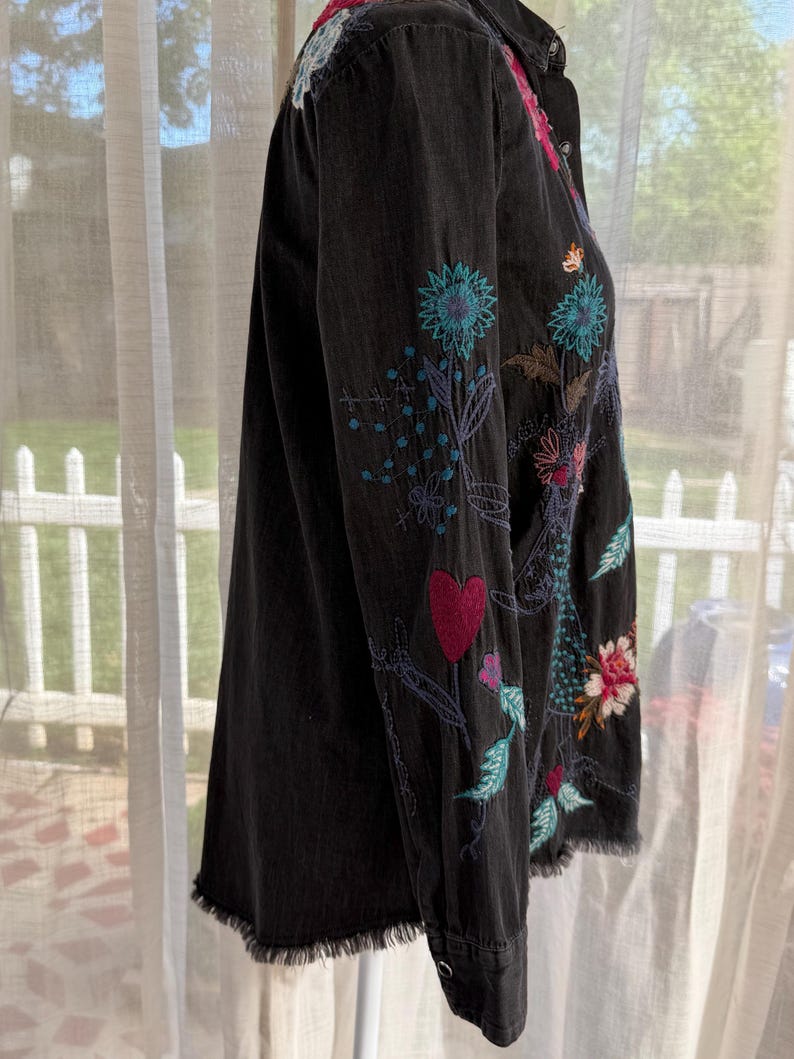 以下が含まれることがあります： カラフルな花の刺繍が施された黒のデニムシャツ。 シャツは、ほつれた裾と長袖が特徴です。 刺繍には、ピンク、青、緑の色合いの花、葉、ハートのデザインが含まれています。