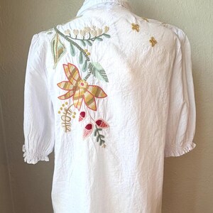 May include: White blouse with floral embroidery on the back. The embroidery features a large flower with yellow, orange, and green colors, along with leaves and small red accents. The sleeves are short and gathered.