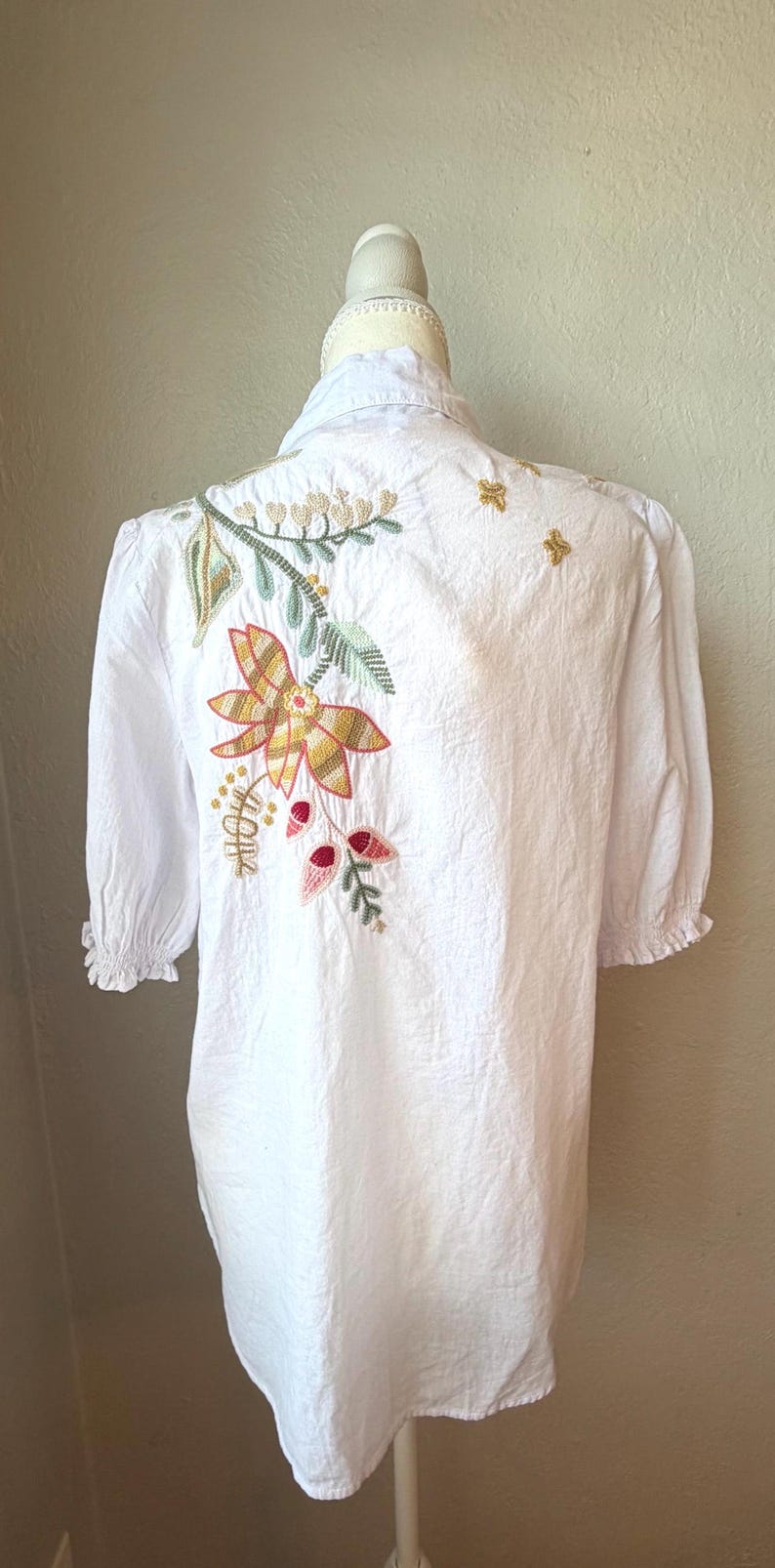 May include: White blouse with floral embroidery on the back. The embroidery features a large flower with yellow, orange, and green colors, along with leaves and small red accents. The sleeves are short and gathered.