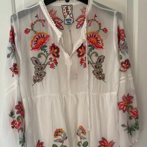 May include: White embroidered tunic dress with long sleeves. The dress features colorful floral embroidery in red, orange, yellow, and green. The neckline and sleeves have delicate trim.