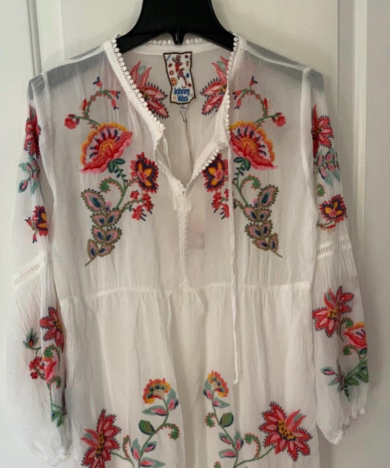May include: White embroidered tunic dress with long sleeves. The dress features colorful floral embroidery in red, orange, yellow, and green. The neckline and sleeves have delicate trim.