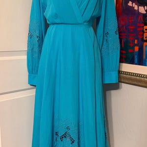 Puede incluir: Un vestido cruzado turquesa con mangas largas y una falda fluida. Las mangas presentan calados florales bordados, y la falda tiene detalles similares cerca del dobladillo. El vestido tiene un escote en V y una cintura definida. Prenda de inspiraci&oacute;n vintage.