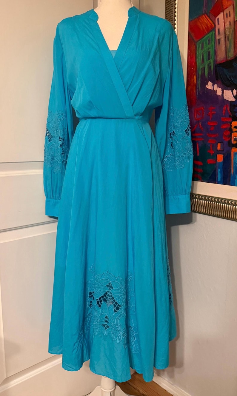 Puede incluir: Un vestido cruzado turquesa con mangas largas y una falda fluida. Las mangas presentan calados florales bordados, y la falda tiene detalles similares cerca del dobladillo. El vestido tiene un escote en V y una cintura definida. Prenda de inspiraci&oacute;n vintage.