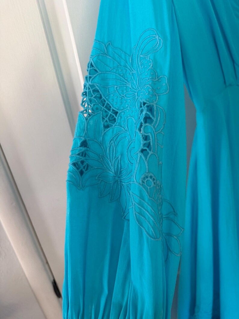 Puede incluir: Vestido turquesa de manga larga con intrincado bordado floral en la manga. El bordado presenta un dise&ntilde;o calado, a&ntilde;adiendo un toque delicado a la prenda. El vestido tiene una silueta fluida.