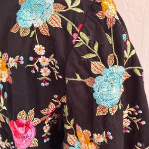 May include: Black jacket with colorful floral embroidery. The embroidery features blue, pink, yellow, and white flowers with green leaves. The jacket's design includes a collar and sleeves, showcasing the detailed floral pattern.