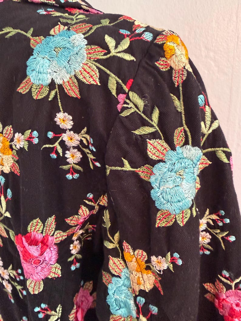 May include: Black jacket with colorful floral embroidery. The embroidery features blue, pink, yellow, and white flowers with green leaves. The jacket's design includes a collar and sleeves, showcasing the detailed floral pattern.