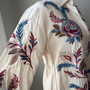 May include: Cream-colored button-down shirt with long sleeves, featuring intricate floral embroidery in shades of blue, burgundy, and brown. The embroidery is concentrated on the sleeves and upper chest, creating a detailed and artistic design.