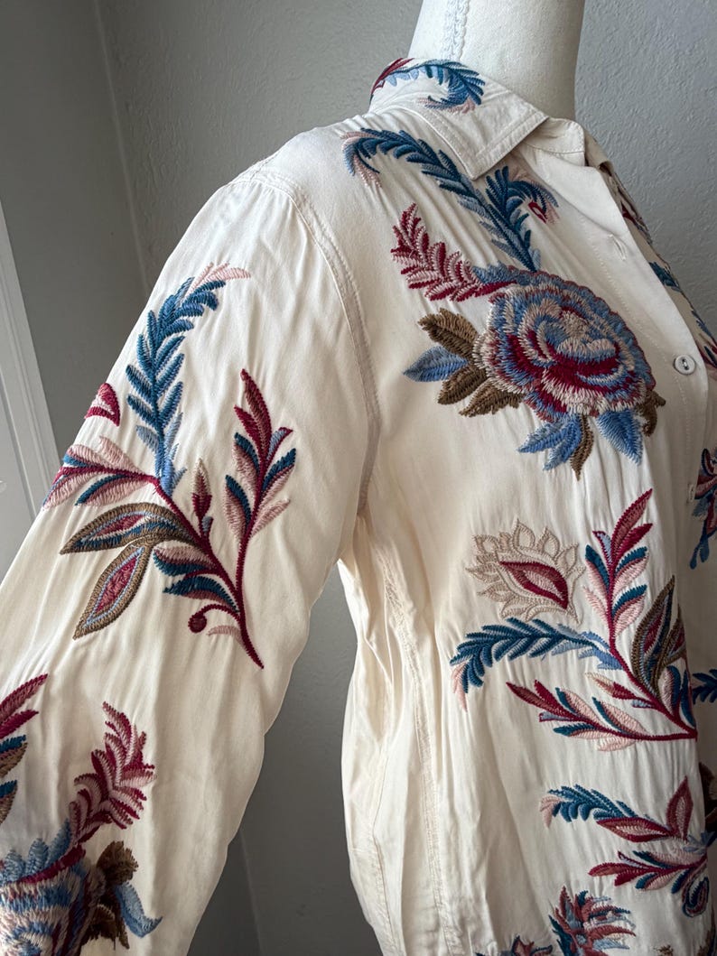 May include: Cream-colored button-down shirt with long sleeves, featuring intricate floral embroidery in shades of blue, burgundy, and brown. The embroidery is concentrated on the sleeves and upper chest, creating a detailed and artistic design.