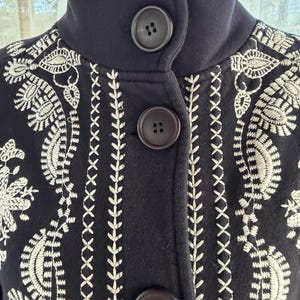 K&ouml;nnte beinhalten: Eine marineblaue Jacke mit hohem Kragen und drei gro&szlig;en, dunkelbraunen Kn&ouml;pfen. Die Jacke hat aufwendige wei&szlig;e Stickereien mit floralen und geometrischen Mustern. Die Stickerei ist detailliert und bedeckt die Vorderseite und die Schultern der Jacke.