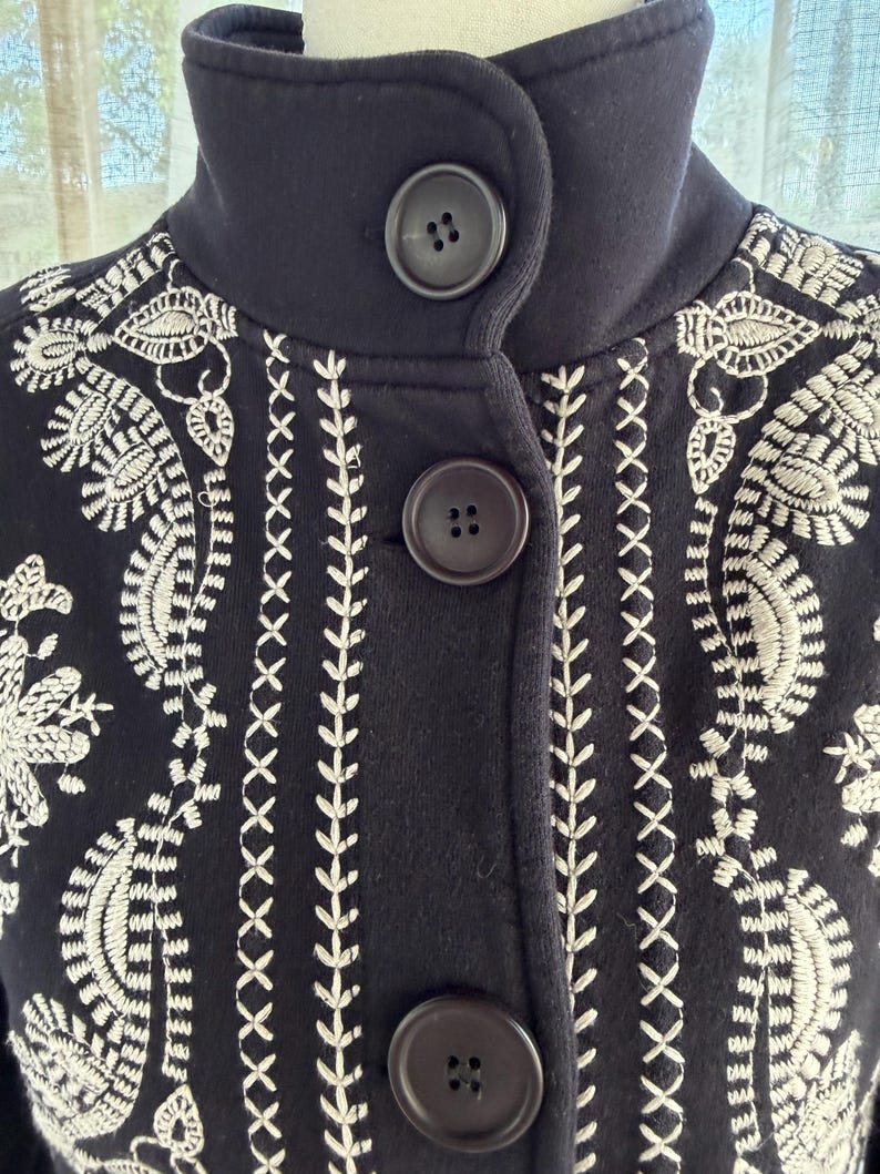 K&ouml;nnte beinhalten: Eine marineblaue Jacke mit hohem Kragen und drei gro&szlig;en, dunkelbraunen Kn&ouml;pfen. Die Jacke hat aufwendige wei&szlig;e Stickereien mit floralen und geometrischen Mustern. Die Stickerei ist detailliert und bedeckt die Vorderseite und die Schultern der Jacke.