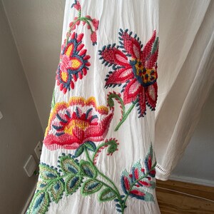 May include: White fabric with vibrant floral embroidery in red, pink, yellow, green, and blue. The embroidered flowers and leaves are detailed and colorful, creating a striking visual contrast against the white background.
