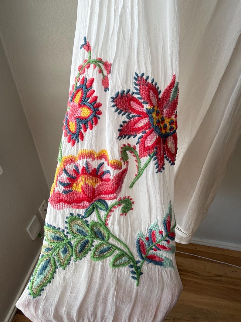 May include: White fabric with vibrant floral embroidery in red, pink, yellow, green, and blue. The embroidered flowers and leaves are detailed and colorful, creating a striking visual contrast against the white background.