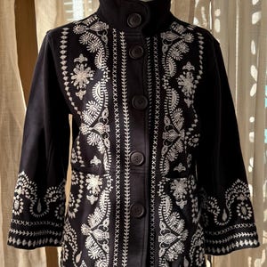 K&ouml;nnte beinhalten: Schwarze Jacke mit wei&szlig;en Stickereien mit floralen und geometrischen Mustern. Die Jacke hat einen hohen Kragen, einen Knopfverschluss und Dreiviertel&auml;rmel. Die Stickerei ist detailliert und bedeckt die Vorderseite, die &Auml;rmel und den Kragen.