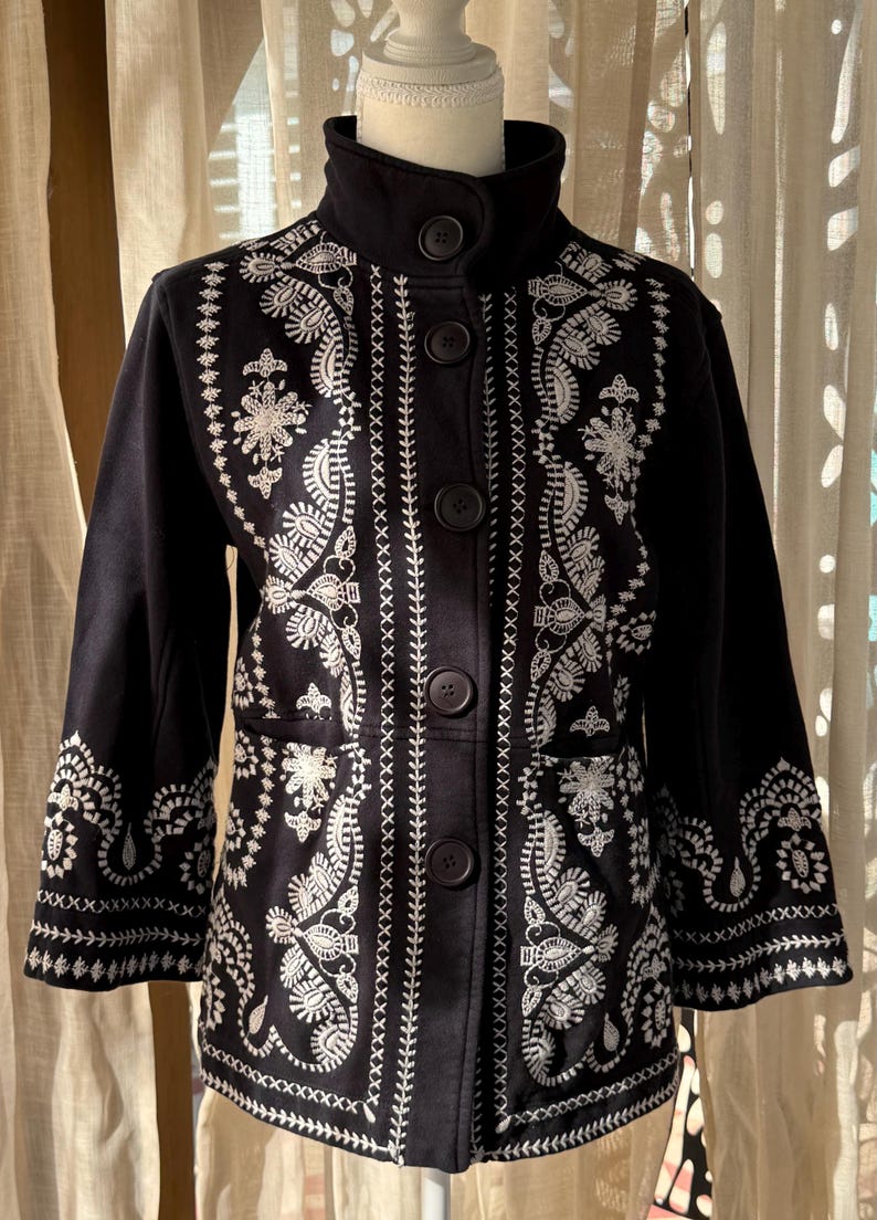 K&ouml;nnte beinhalten: Schwarze Jacke mit wei&szlig;en Stickereien mit floralen und geometrischen Mustern. Die Jacke hat einen hohen Kragen, einen Knopfverschluss und Dreiviertel&auml;rmel. Die Stickerei ist detailliert und bedeckt die Vorderseite, die &Auml;rmel und den Kragen.