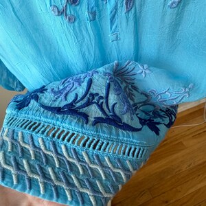 Puede incluir: T&uacute;nica azul claro bordada con intrincados patrones florales y geom&eacute;tricos. La t&uacute;nica presenta un cuello mandar&iacute;n y mangas largas con bordados detallados en los pu&ntilde;os. La tela tiene una textura ligeramente arrugada.
