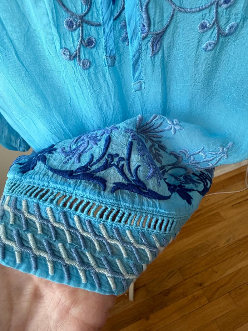 Puede incluir: T&uacute;nica azul claro bordada con intrincados patrones florales y geom&eacute;tricos. La t&uacute;nica presenta un cuello mandar&iacute;n y mangas largas con bordados detallados en los pu&ntilde;os. La tela tiene una textura ligeramente arrugada.