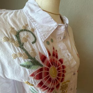 May include: White button-down shirt with colorful floral embroidery. The embroidery features a large red and yellow flower, green leaves, and other floral accents. The shirt is displayed on a mannequin.