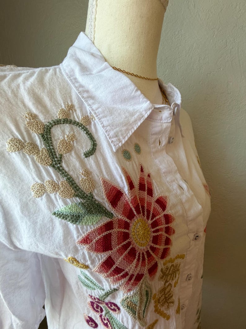 May include: White button-down shirt with colorful floral embroidery. The embroidery features a large red and yellow flower, green leaves, and other floral accents. The shirt is displayed on a mannequin.