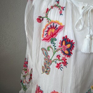 May include: White blouse with long sleeves and colorful floral embroidery. The embroidery features red, pink, yellow, and blue flowers and leaves. The blouse has a tie-neck closure with white tassels.