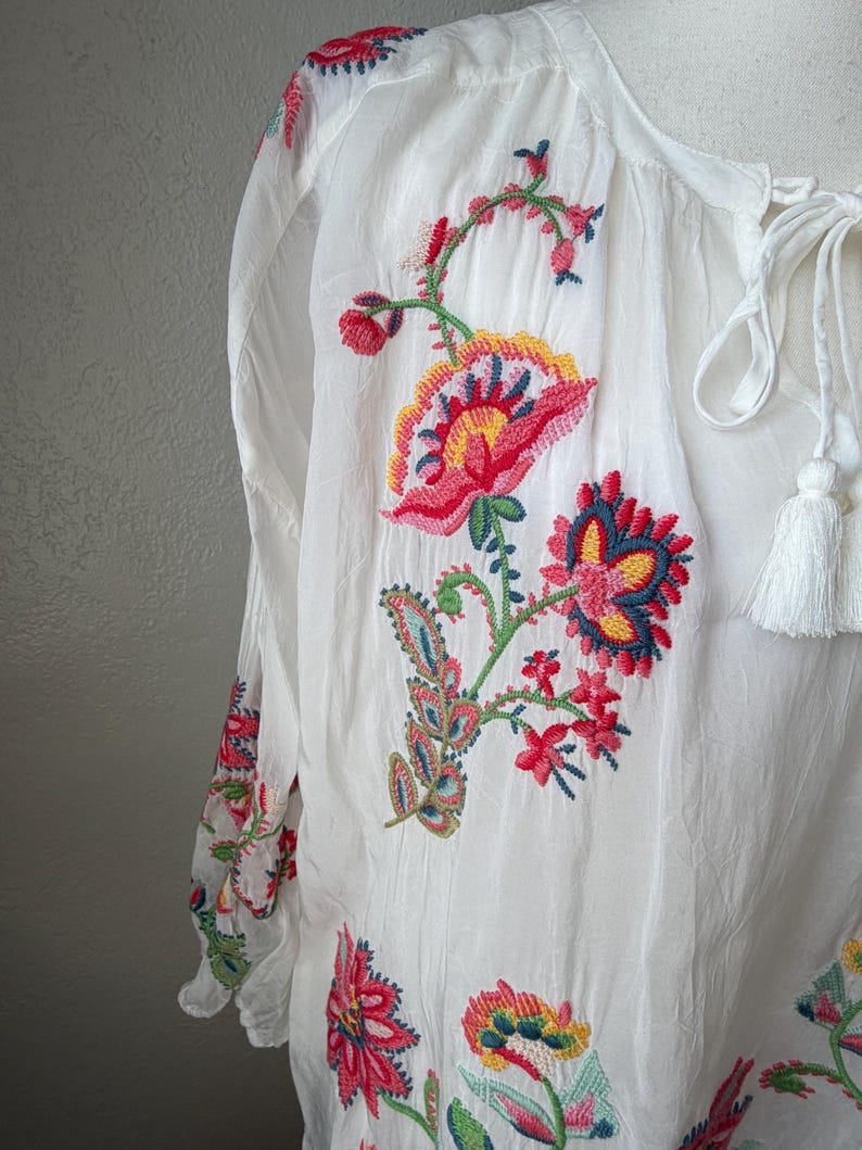 May include: White blouse with long sleeves and colorful floral embroidery. The embroidery features red, pink, yellow, and blue flowers and leaves. The blouse has a tie-neck closure with white tassels.