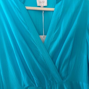 Puede incluir: Vestido turquesa con cuello en V y mangas largas, colgado de una percha de madera. La etiqueta dice "jade". La tela parece ser un material ligero y fluido. El vestido es de un tono azul vibrante.