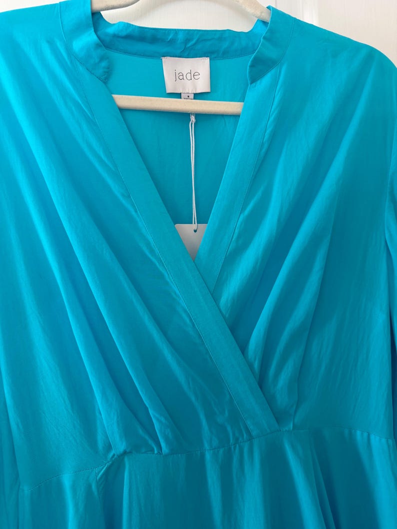 Puede incluir: Vestido turquesa con cuello en V y mangas largas, colgado de una percha de madera. La etiqueta dice "jade". La tela parece ser un material ligero y fluido. El vestido es de un tono azul vibrante.