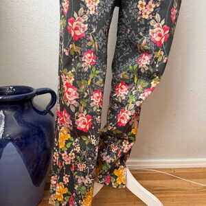 Puede incluir: Un pantal&oacute;n gris oscuro con un vibrante estampado floral de rosas rojas, rosas y amarillas, hojas verdes y un sutil fondo de damasco negro. El pantal&oacute;n se muestra sobre un soporte blanco, exhibiendo el dise&ntilde;o de pierna larga.